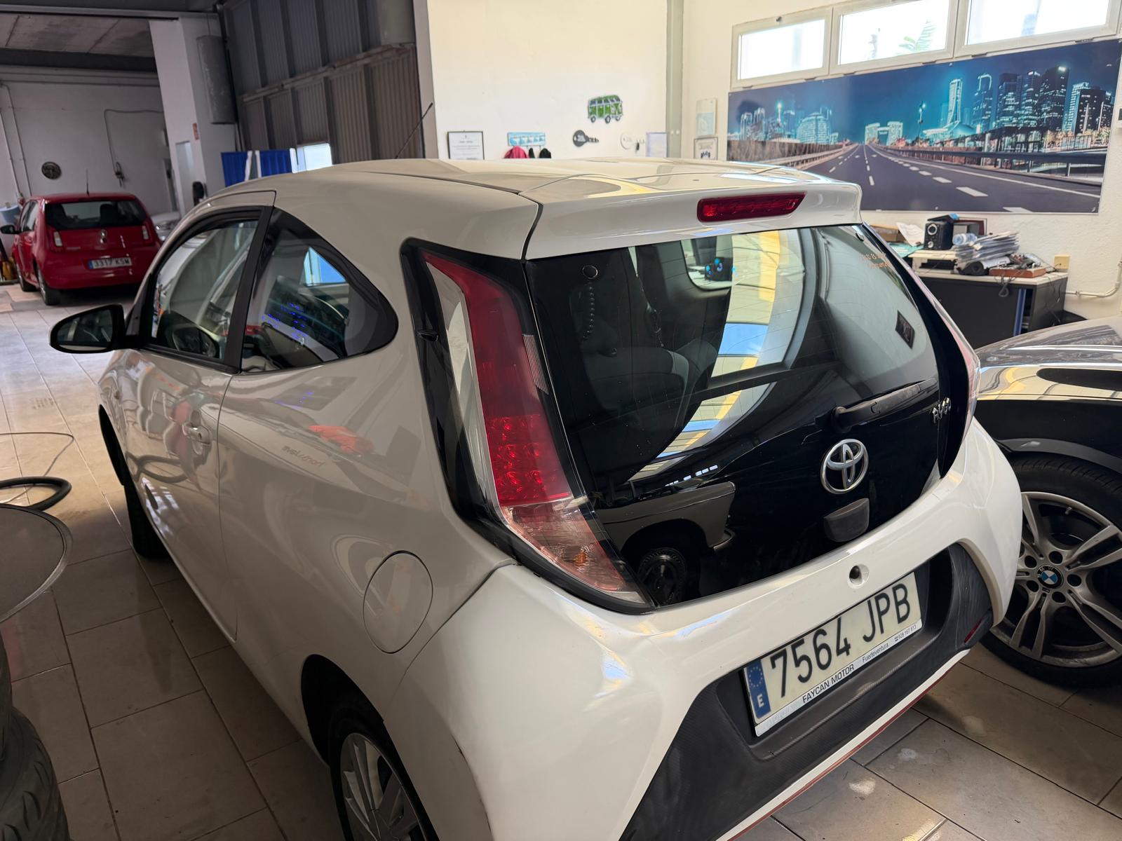 Toyota Aygo blanco estacionado en un taller mecánico, con vista lateral trasera que muestra su diseño compacto y moderno. Al fondo, se pueden ver otros vehículos, incluyendo un coche rojo.