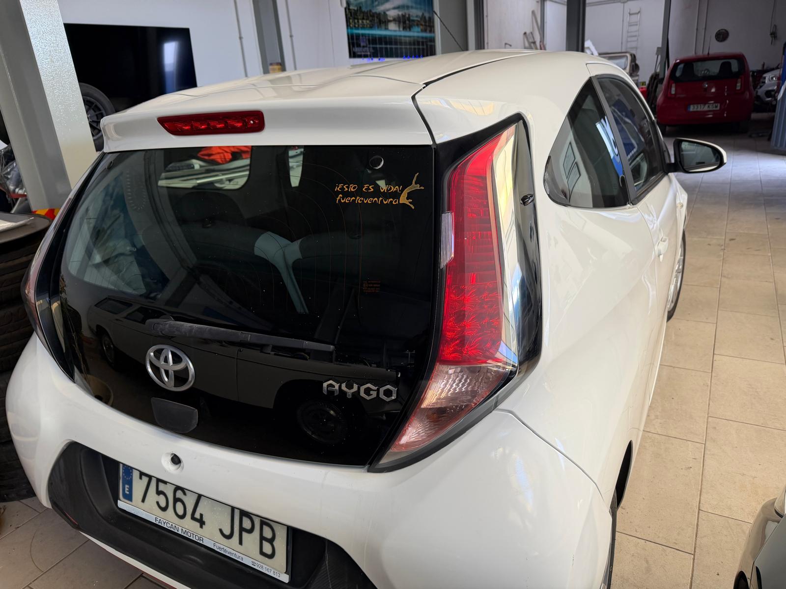 Vista trasera de un Toyota Aygo blanco estacionado en un taller, mostrando el logo de la marca y detalles del diseño del vehículo.