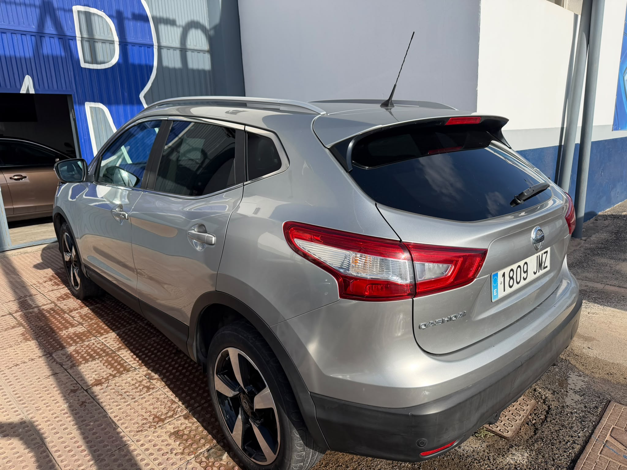 SUV Nissan Qashqai de color plata estacionado frente a un taller, con detalles visibles de su diseño moderno y llantas deportivas.