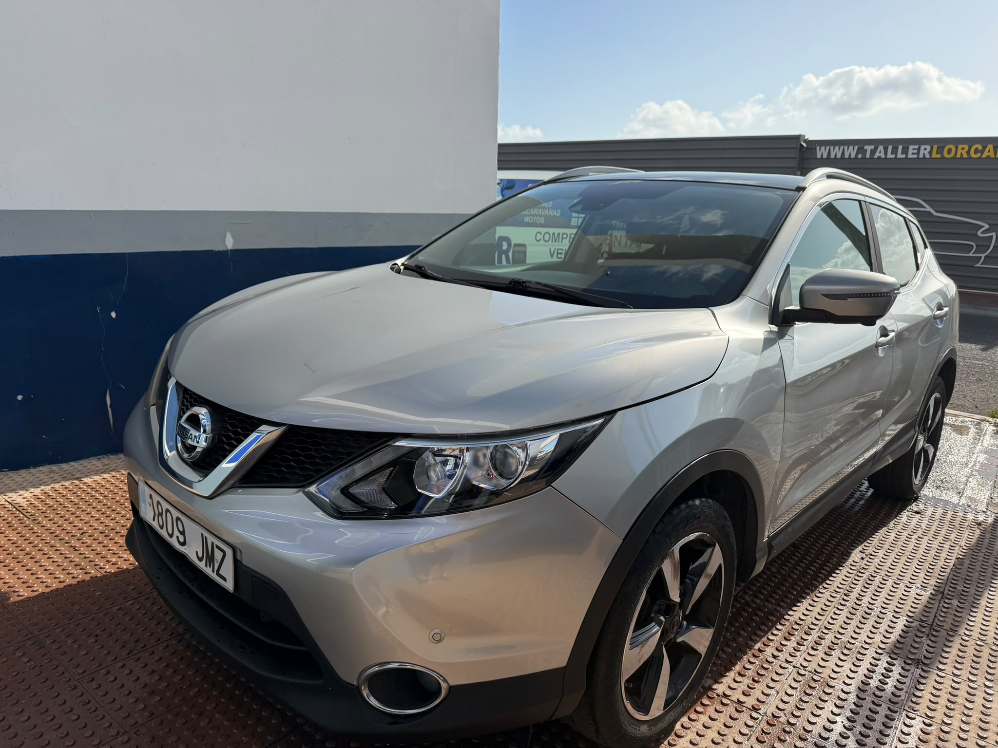 SUV Nissan en color plata estacionado frente a un taller mecánico, mostrando un diseño moderno y elegante, con llantas de aleación y detalles distintivos en la parrilla.