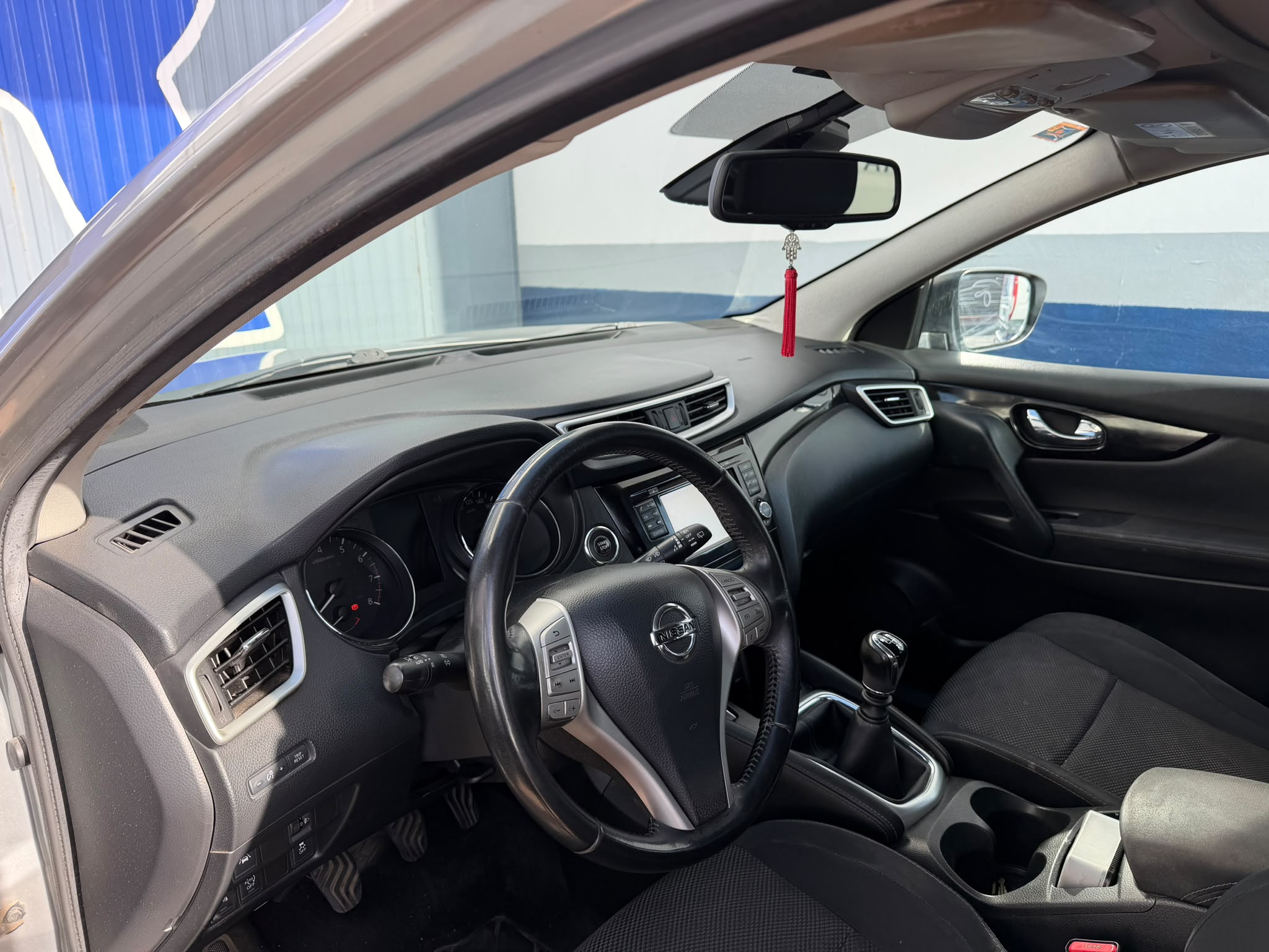 Interior de un automóvil Nissan con volante, tablero de instrumentos y palanca de cambios. Se observa un espejo retrovisor con un adorno, asientos de tela y un diseño moderno. Fondo azul claro visible a través de la ventana.