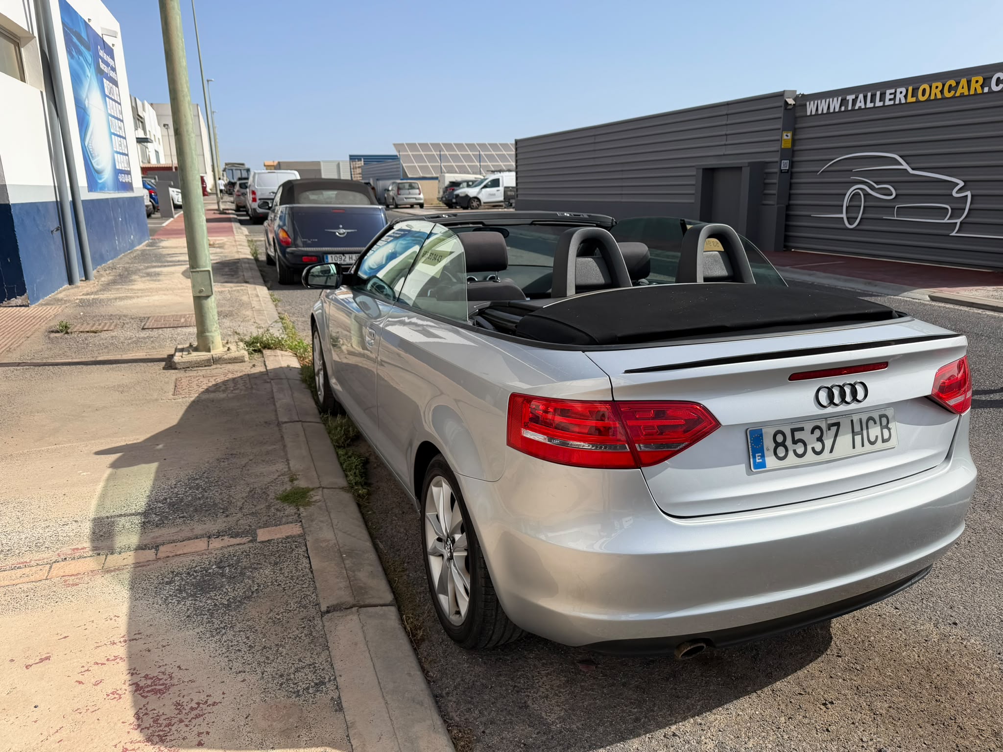 Coche descapotable Audi plateado estacionado en la acera, con un taller de automóviles y otros vehículos de fondo, bajo un cielo despejado.