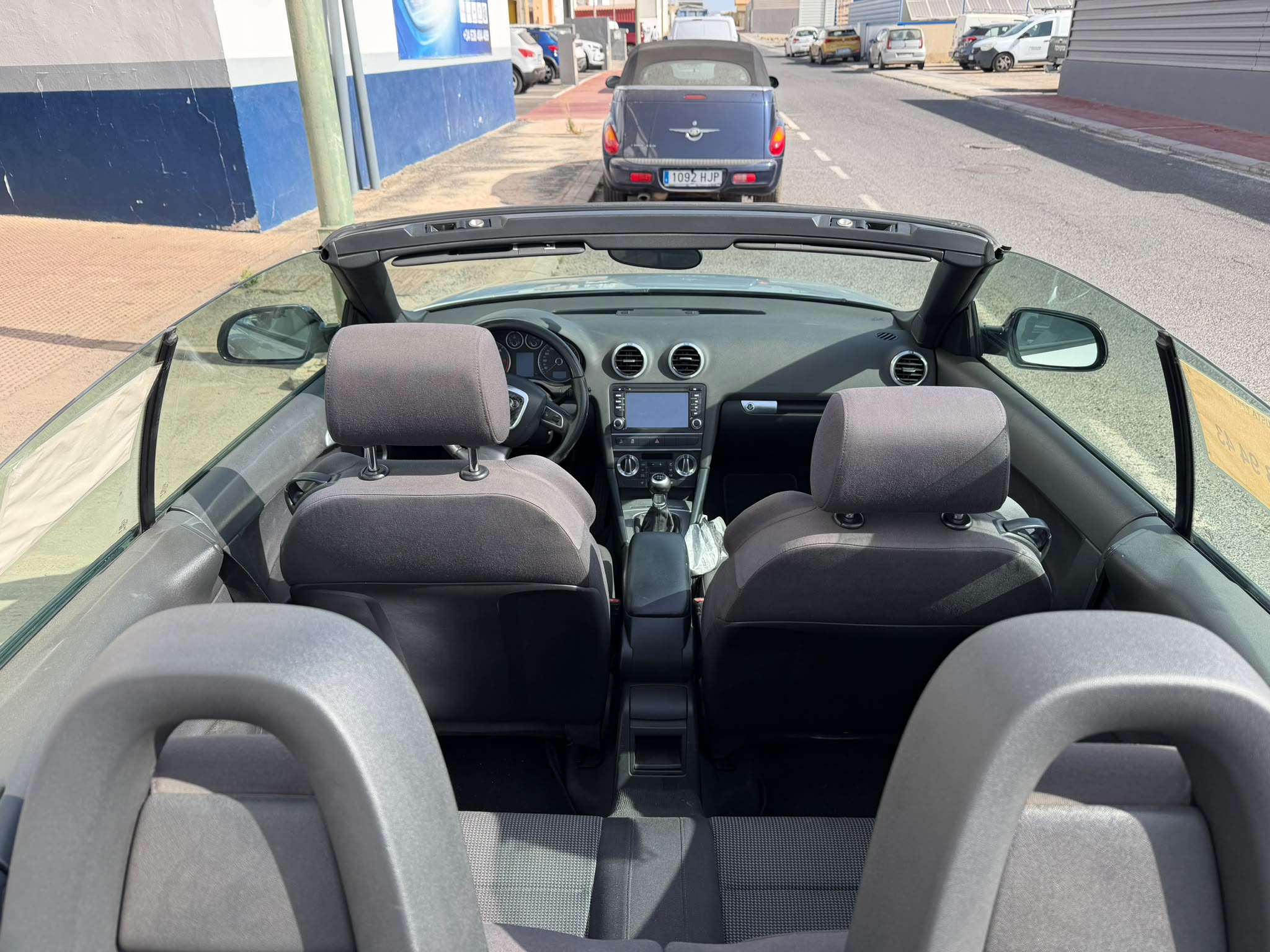 Interior de un coche descapotable, mostrando asientos traseros y el tablero de instrumentos. Se observa la carretera y otros vehículos estacionados en el fondo. Ideal para resaltar la comodidad y diseño de vehículos convertibles.