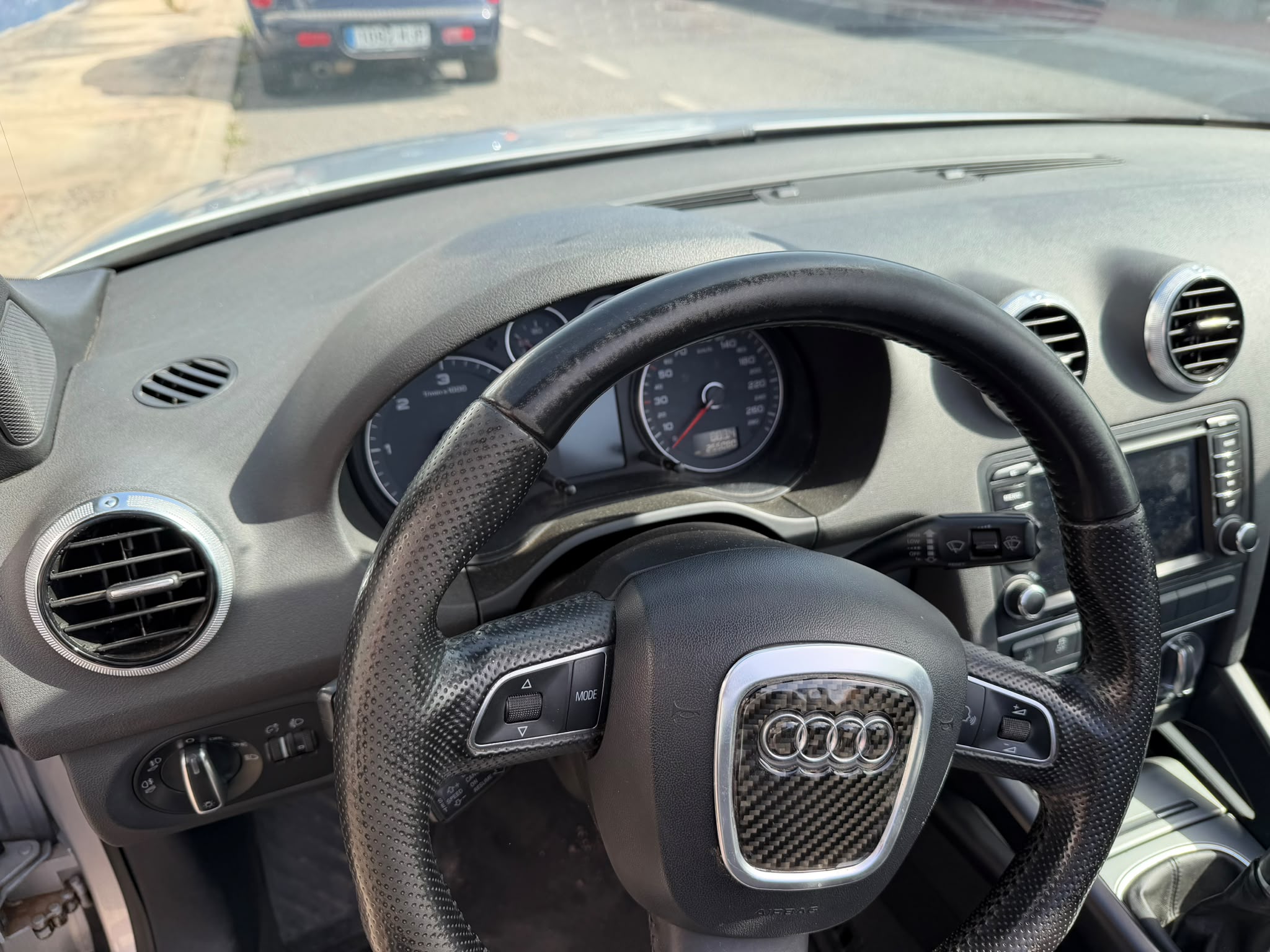 Interior de un automóvil Audi, mostrando el volante y el tablero de instrumentos. Se aprecian los controles del volante, las salidas de aire acondicionado y una pantalla multimedia. El paisaje exterior incluye un vehículo estacionado en la calle.