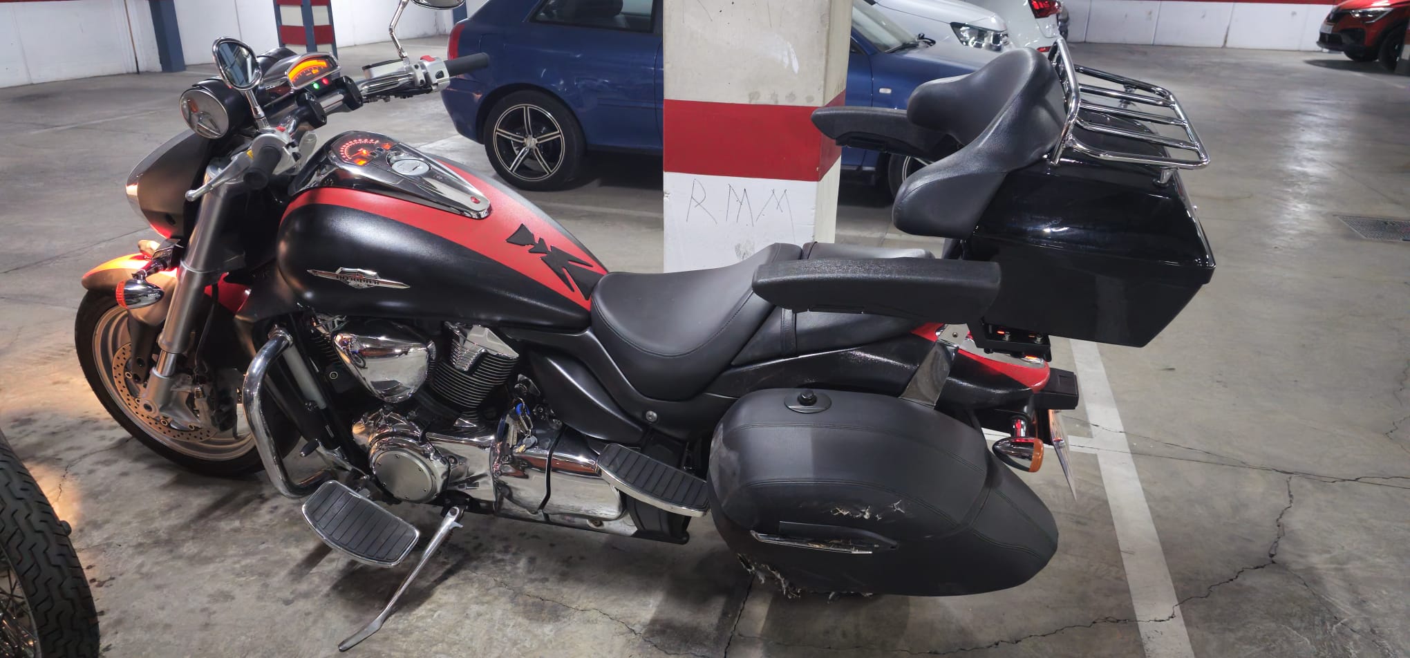 Motocicleta de estilo cruiser con diseño en negro y rojo, estacionada en un garaje. Se destacan sus detalles cromados y maleteros laterales, ideal para viajes largos.