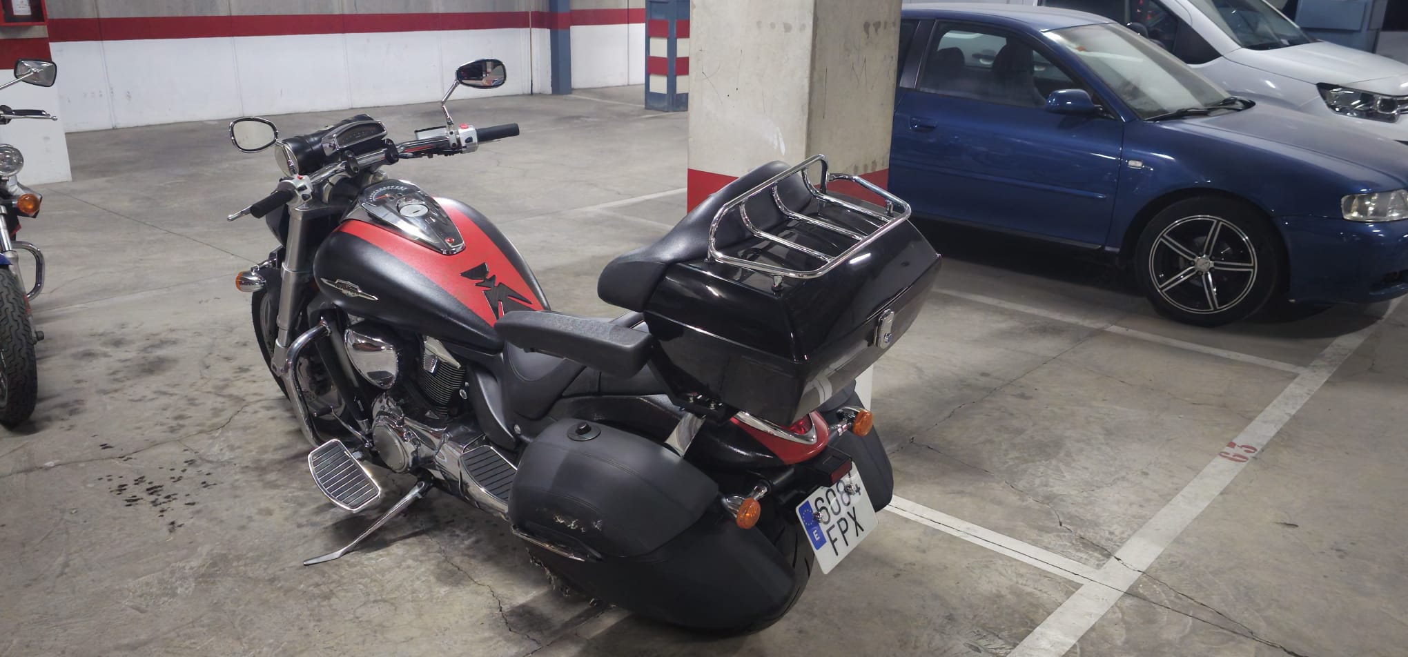 Motocicleta negra con detalles rojos estacionada en un garaje, con un baúl trasero y otras vehículos visibles al fondo.