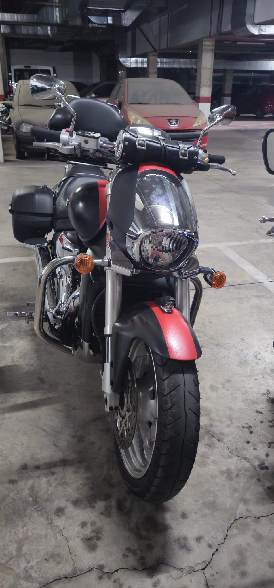 Motocicleta de estilo cruiser en un estacionamiento, con carrocería negra y detalles rojos, equipada con maleteros laterales y faros cromados, destacando sus llantas anchas y diseño robusto.