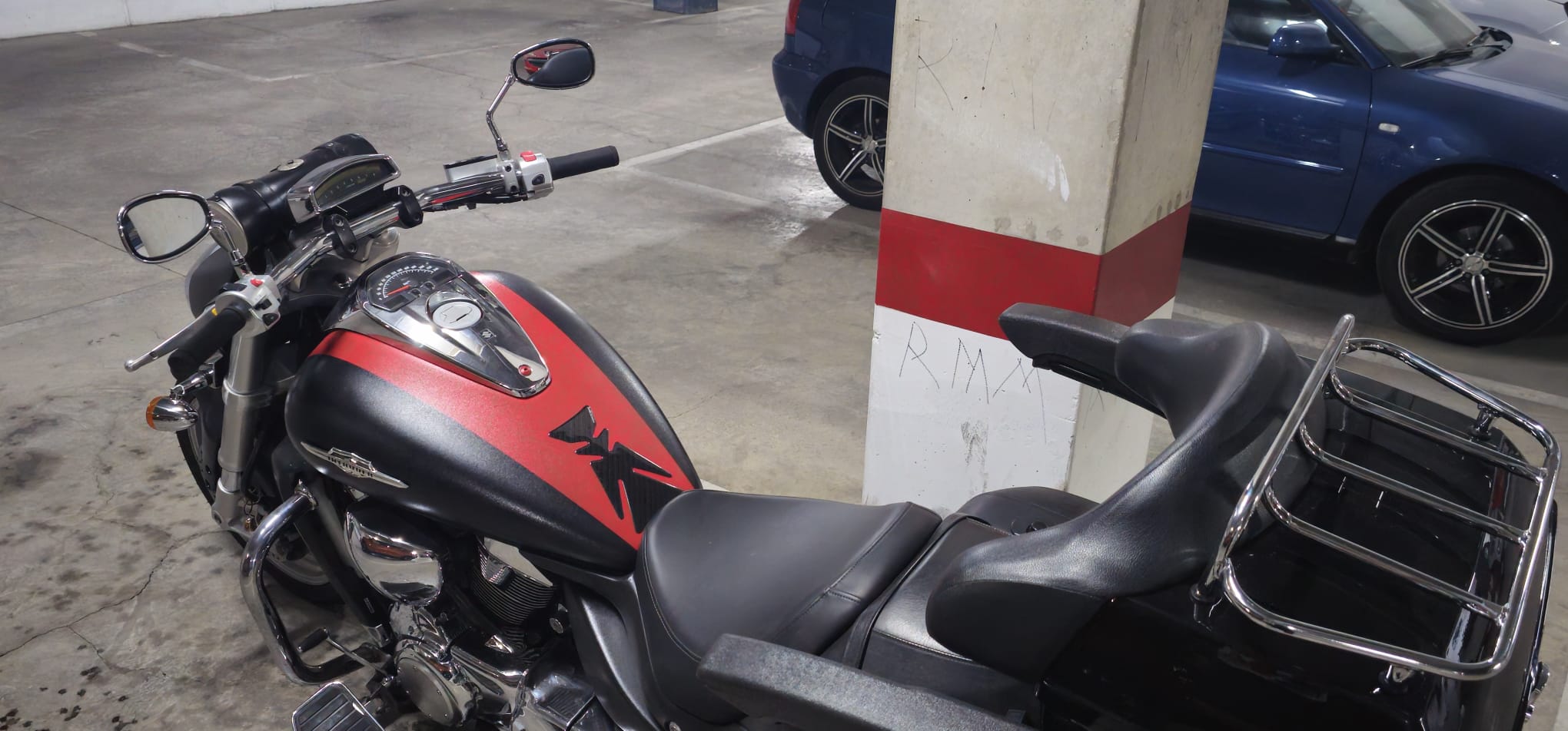 Motocicleta con diseño en rojo y negro estacionada en un aparcamiento, mostrando detalles del manillar y el asiento. Fondo con otros vehículos y columna de soporte.