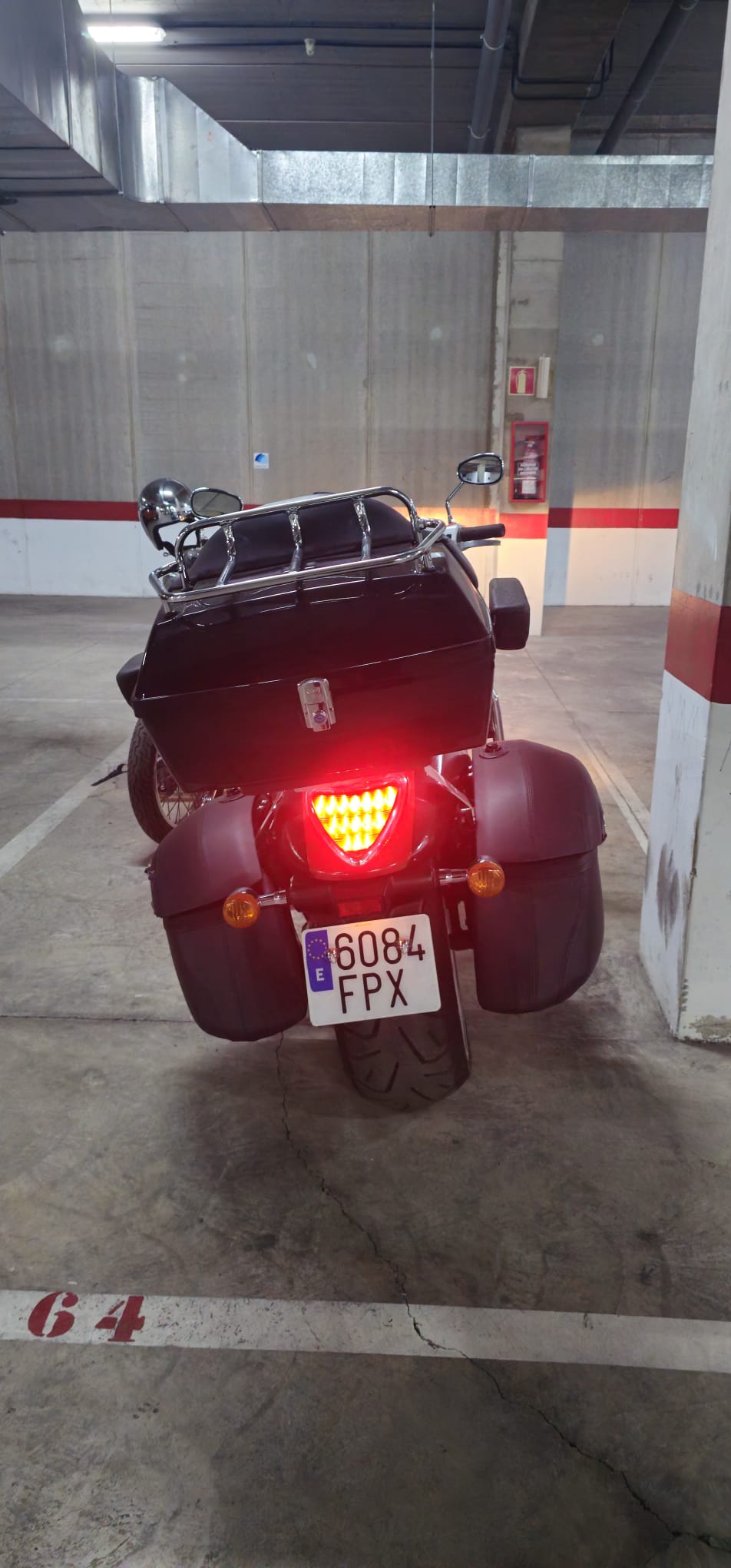 Motocicleta aparcada en un garaje, vista trasera con maletero negro y luces traseras encendidas. Número de matrícula 6084 FPX visible en el lado derecho. Suelo de concreto con marcas de estacionamiento.