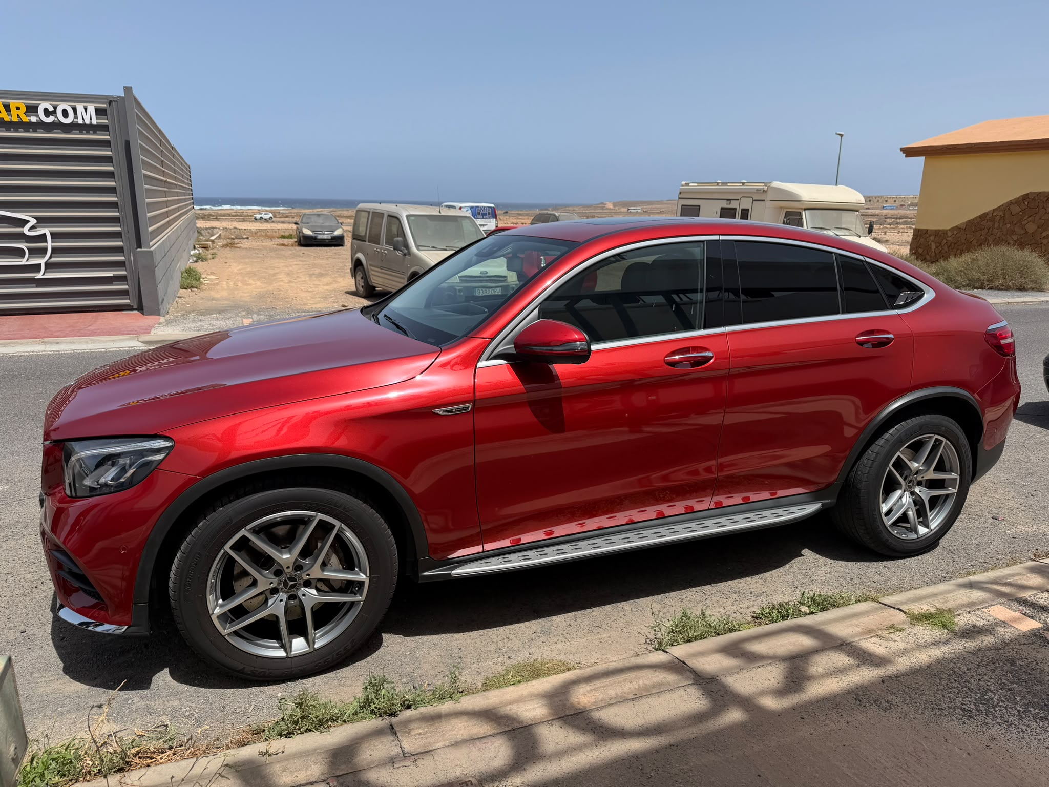 SUV de lujo color rojo aparcado en la calle, con llantas de aleación y un diseño aerodinámico. Al fondo, se pueden ver otros vehículos y un paisaje costero.