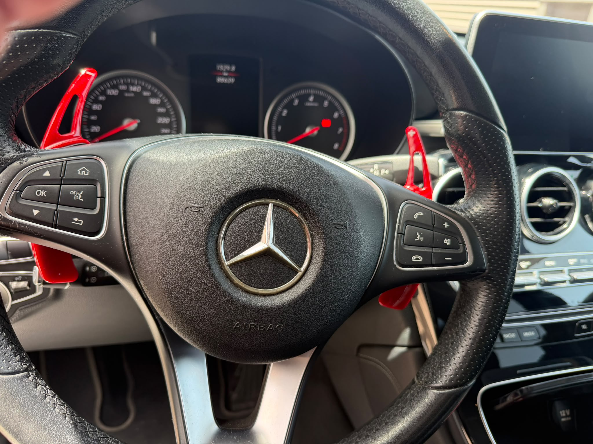 Interior de un automóvil Mercedes-Benz, mostrando el volante con detalles en rojo y controles de audio y teléfono. El tablero de instrumentos y la pantalla central son visibles, destacando un diseño moderno y elegante.