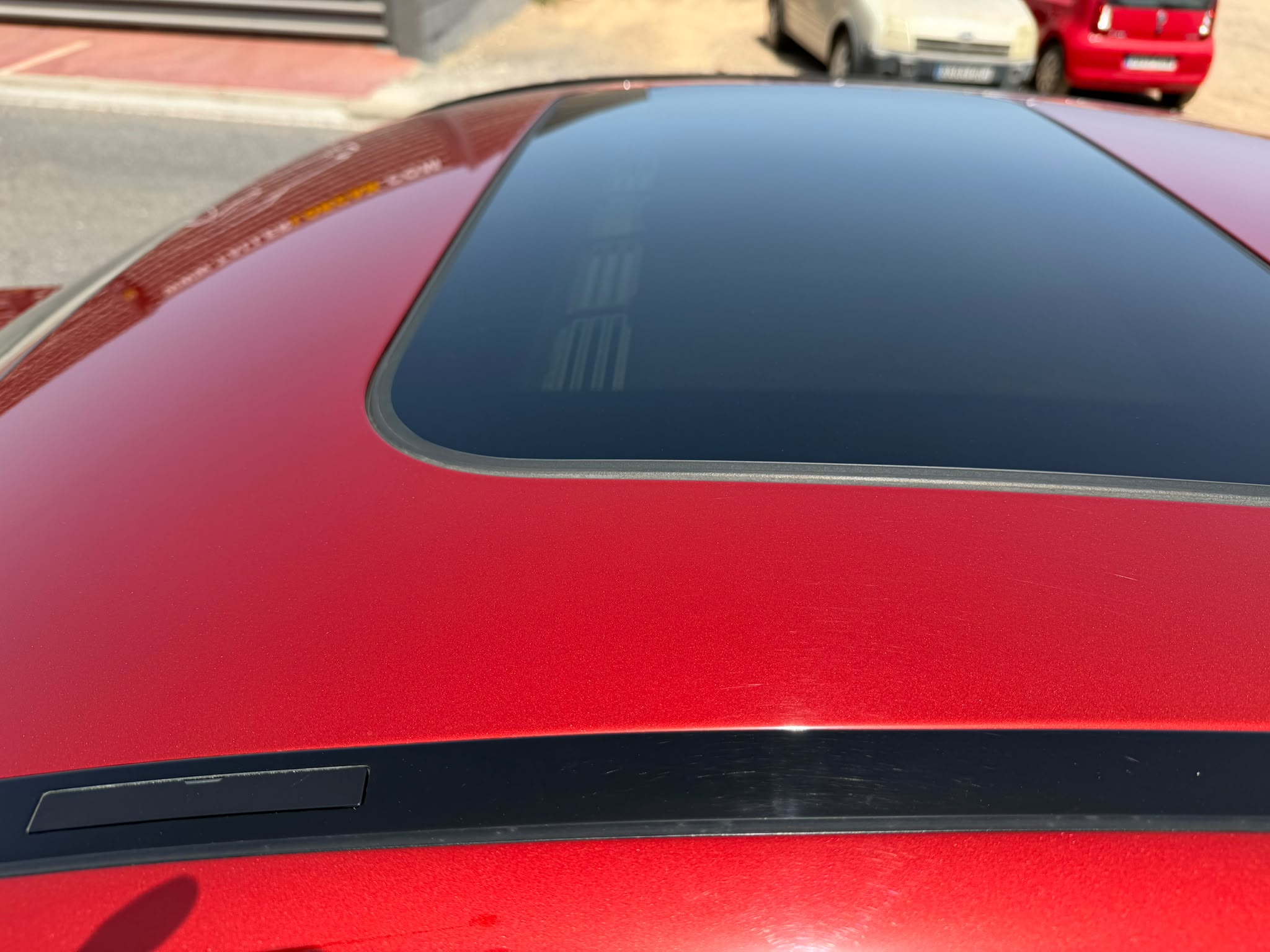 Vista superior del techo de un coche rojo con un techo panorámico. Se observan detalles del acabado y la moldura del vidrio. En el fondo, se pueden ver otros vehículos estacionados.