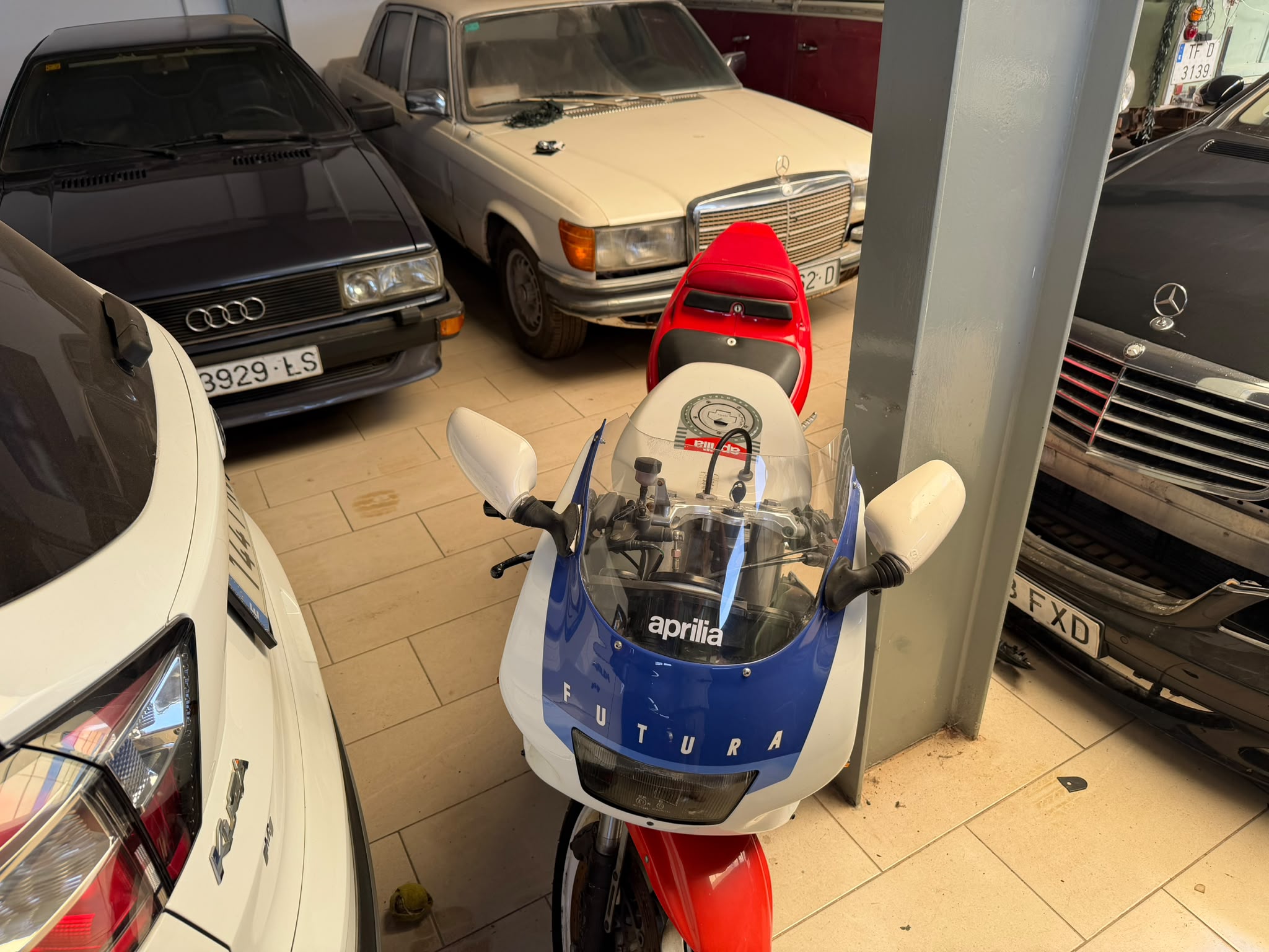 Moto Aprilia Futura en un garaje con varios coches clásicos, incluyendo un Audi y varios modelos de Mercedes-Benz. La imagen muestra una variedad de vehículos estacionados en un espacio reducido.