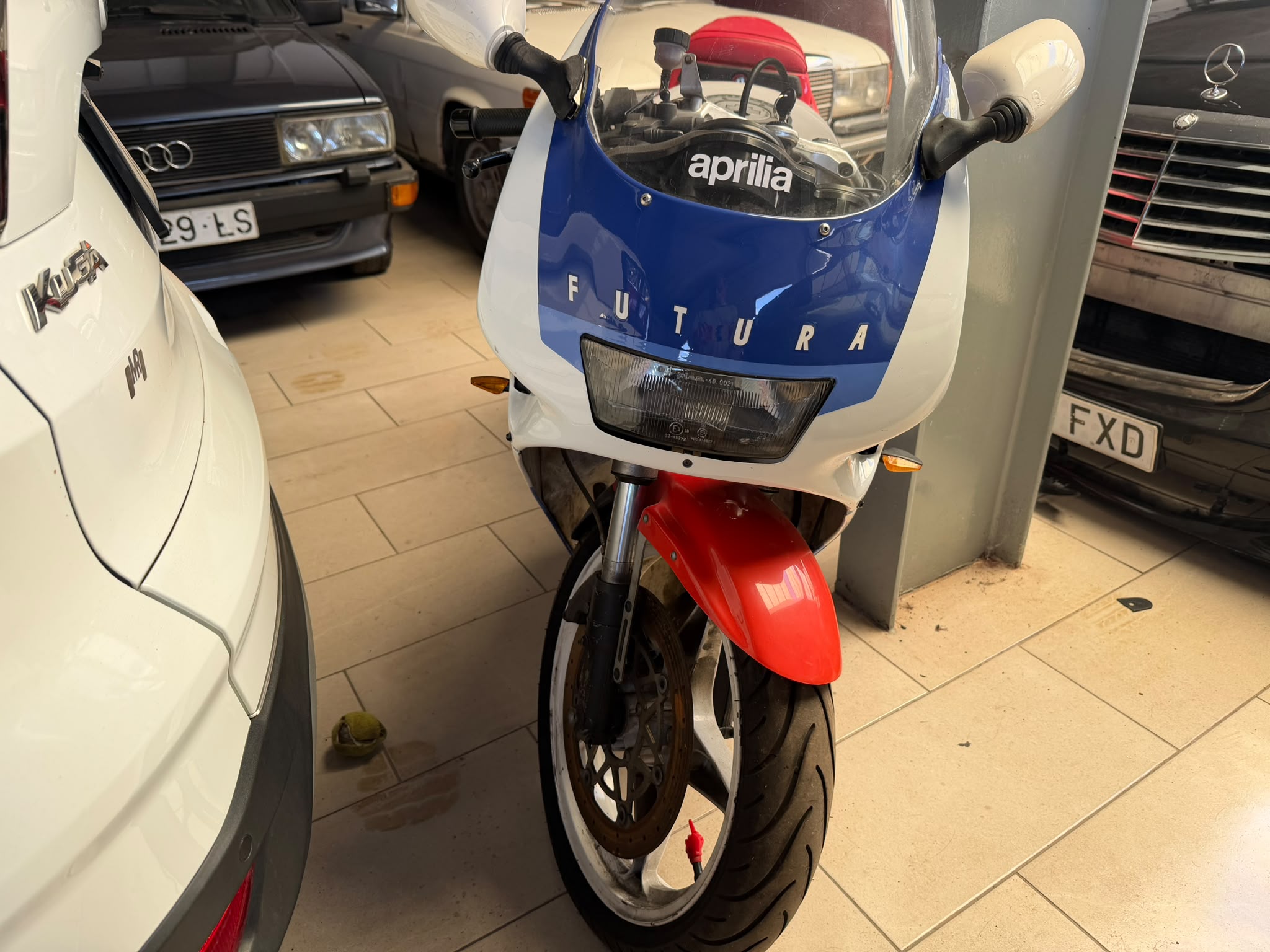 Motocicleta Aprilia Futura en un garaje, rodeada de varios vehículos, incluyendo un Audi y un Mercedes. La moto destaca por su diseño clásico y colores azul y rojo.