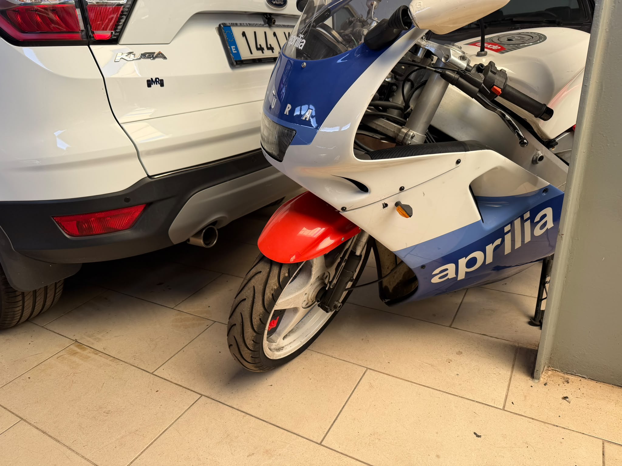 Motocicleta Aprilia estacionada junto a un automóvil Ford Kuga en un garaje, mostrando detalles del diseño y la rueda delantera.