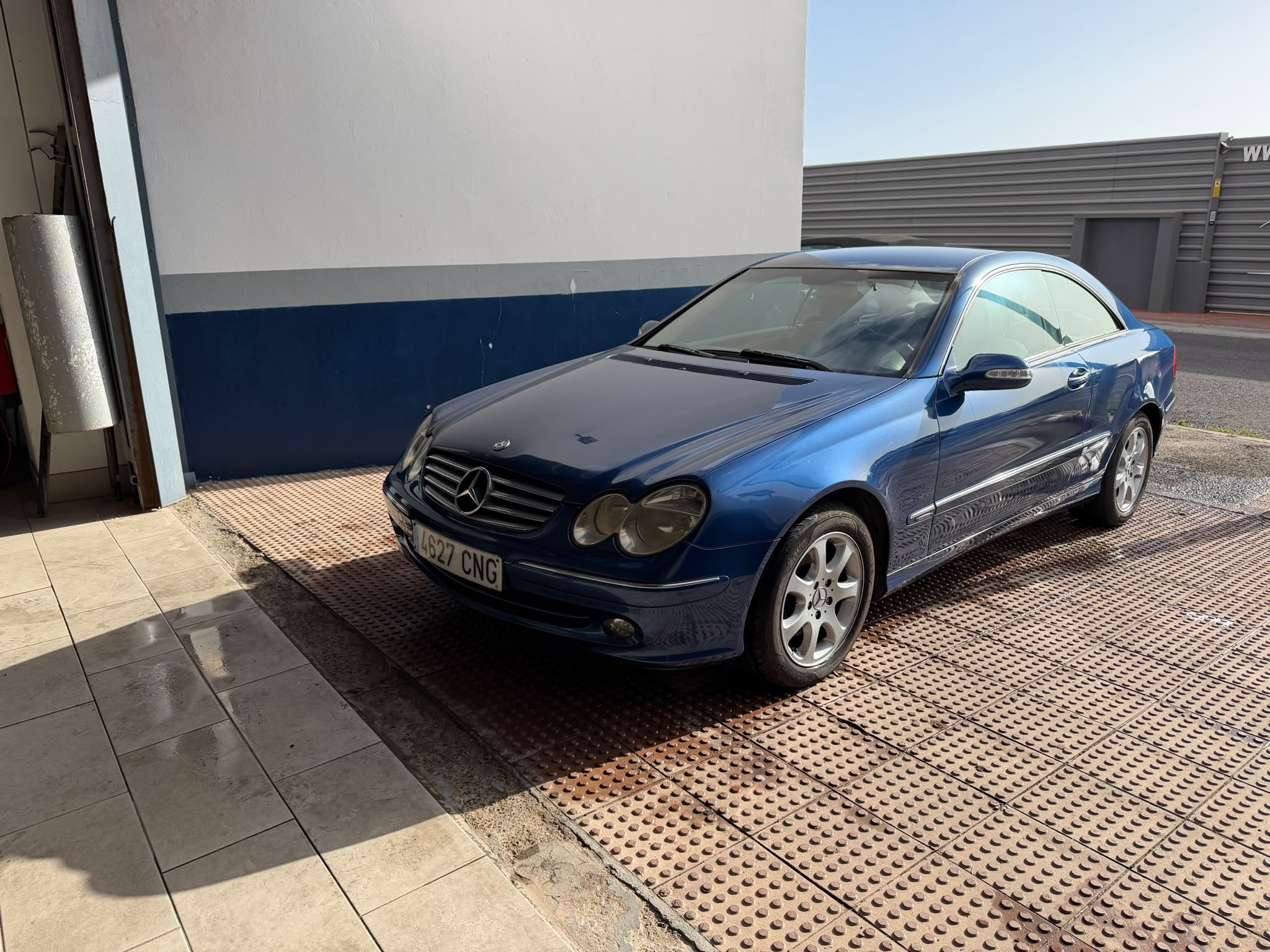 Coche Mercedes-Benz azul estacionado frente a un taller, mostrando su diseño elegante y llantas de aleación, con un fondo de pared blanca y azul.