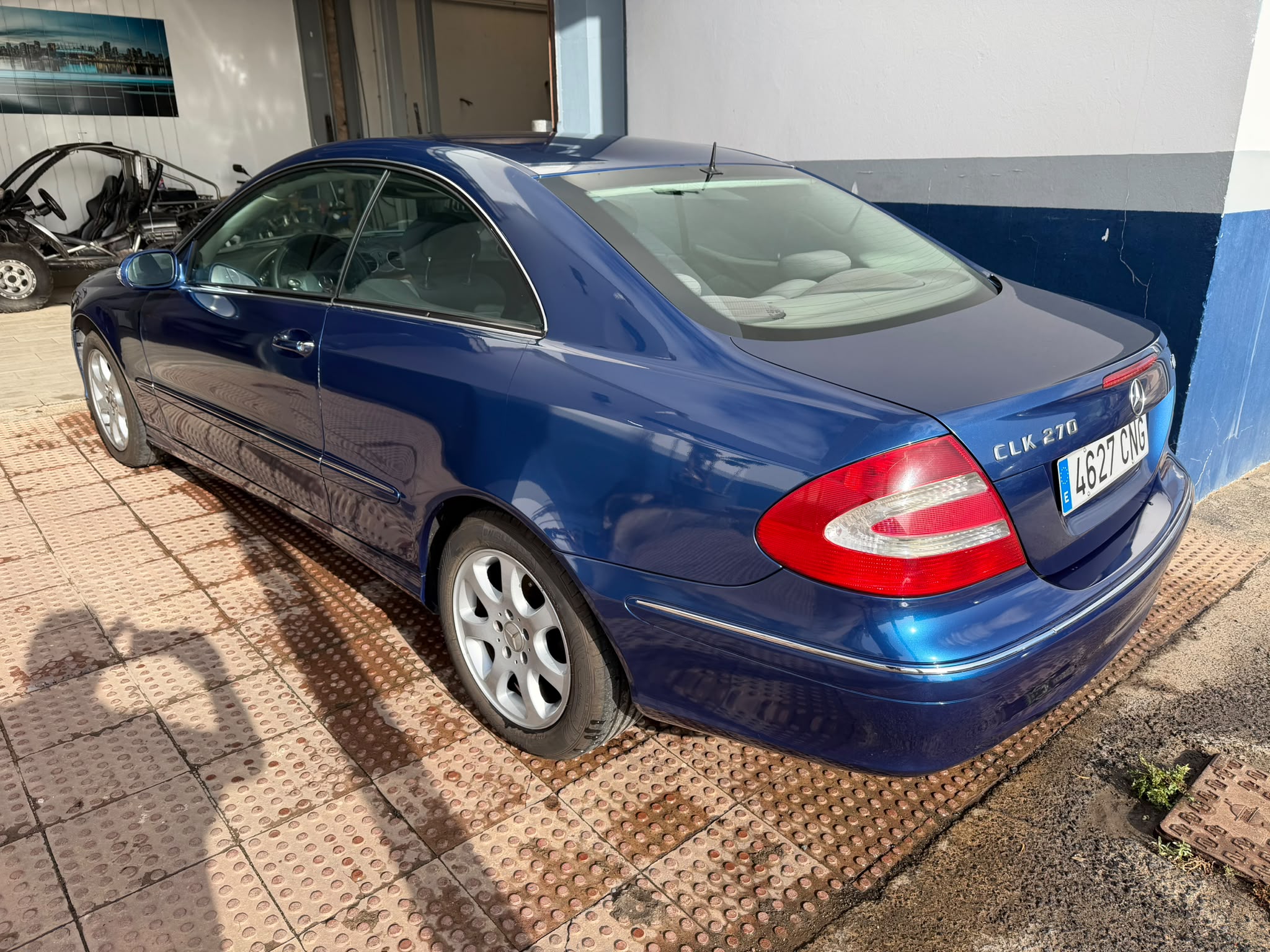 Coche Mercedes-Benz CLK 270 de color azul estacionado, mostrando su diseño elegante y deportivo, con llantas de aleación y un fondo de taller automotriz.