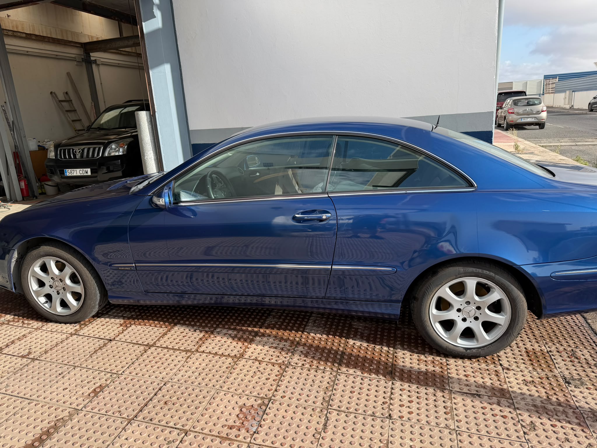 Coche deportivo azul estacionado en un garaje, mostrando su diseño elegante y llantas de aleación. En el fondo, se pueden ver otros vehículos y un entorno de taller.