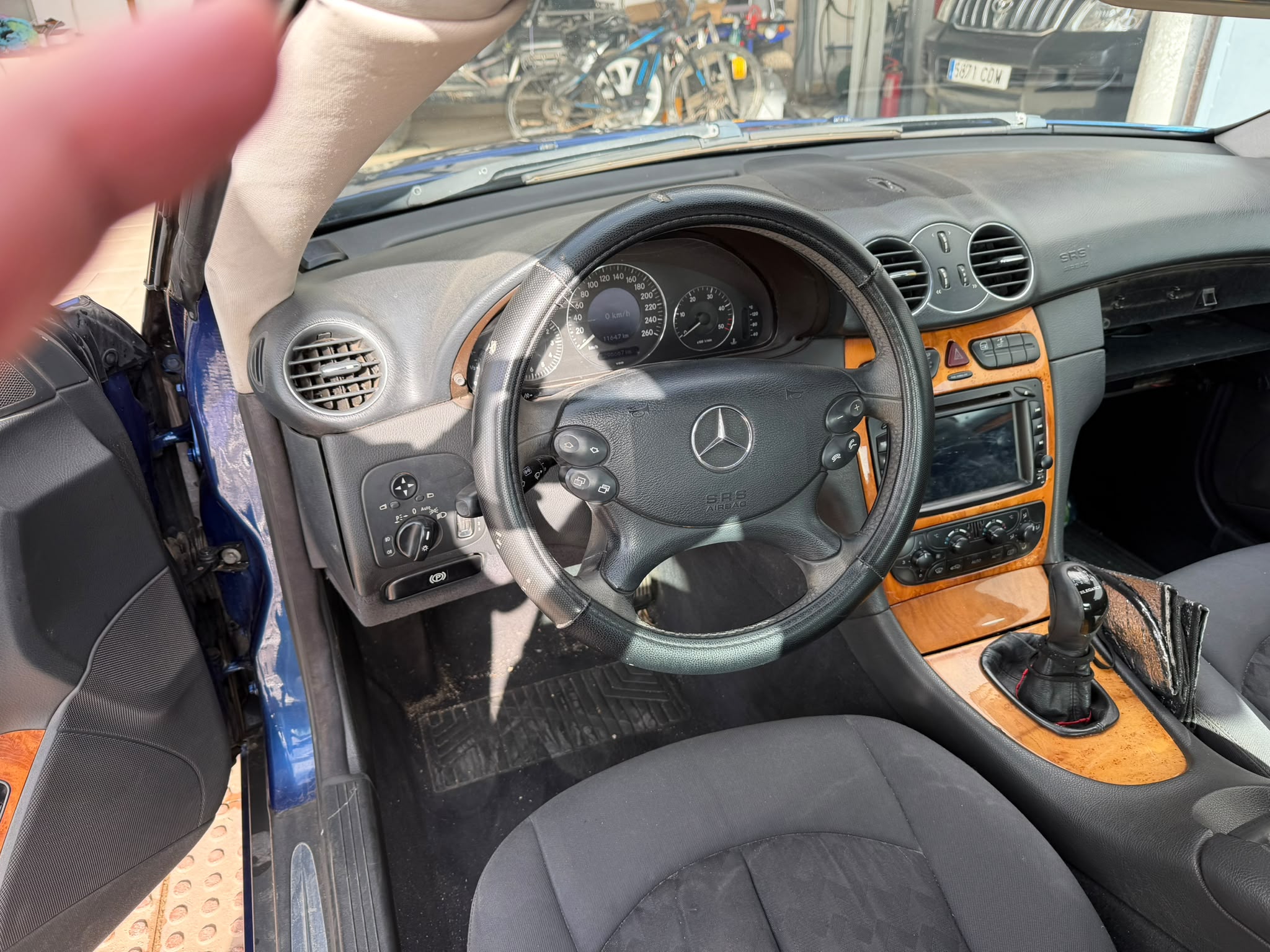 Interior de un automóvil Mercedes-Benz con volante deportivo, panel de instrumentos digital y detalles en madera. Se observa la palanca de cambios y el sistema de audio en la consola central.