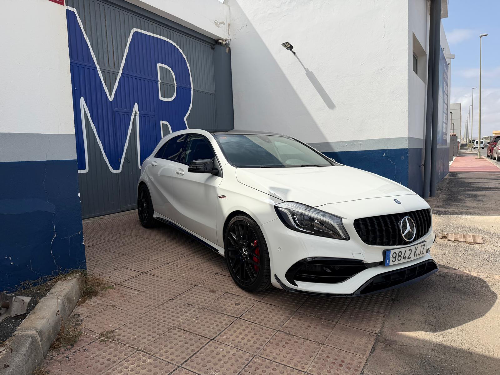 Mercedes-Benz CLA AMG en color blanco estacionado frente a una puerta con el logo "MR". El vehículo destaca por su diseño deportivo y llantas negras, en un entorno urbano.
