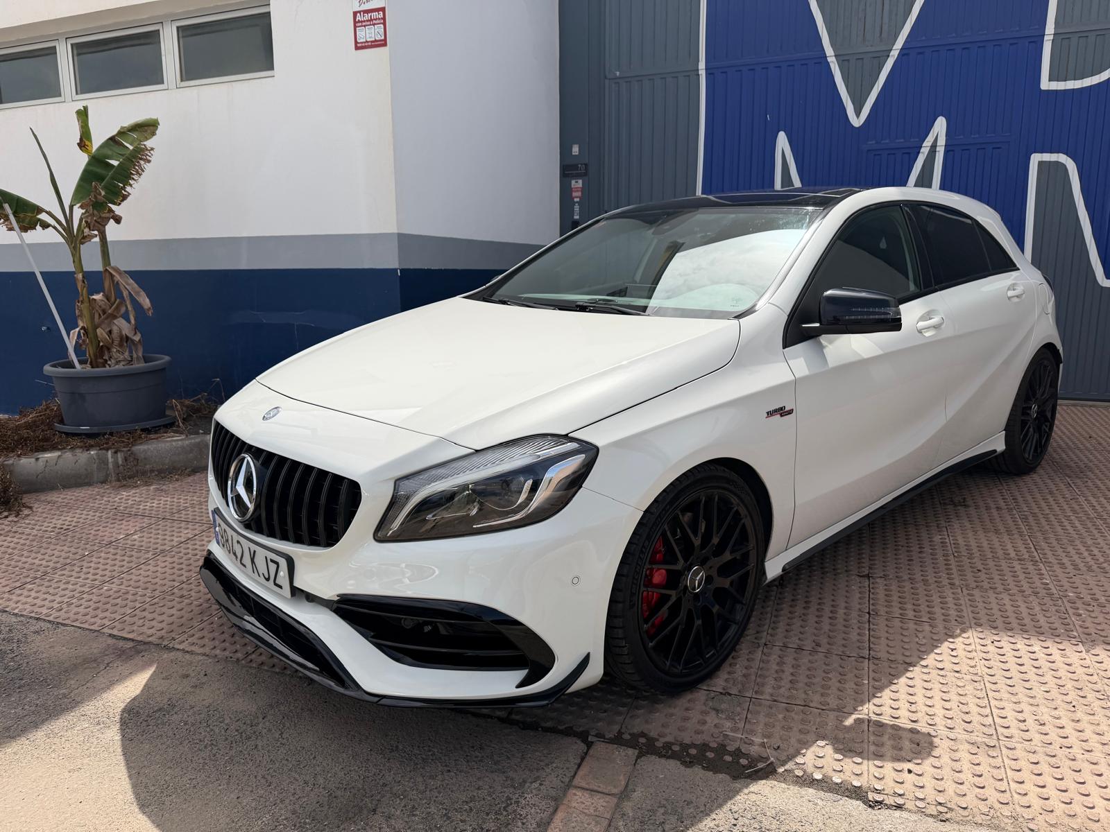 Mercedes-Benz A-Class AMG de color blanco estacionado en un área pavimentada, con llantas de aleación negra y detalles deportivos. Al fondo, se observa una pared azul con el logo de la marca.