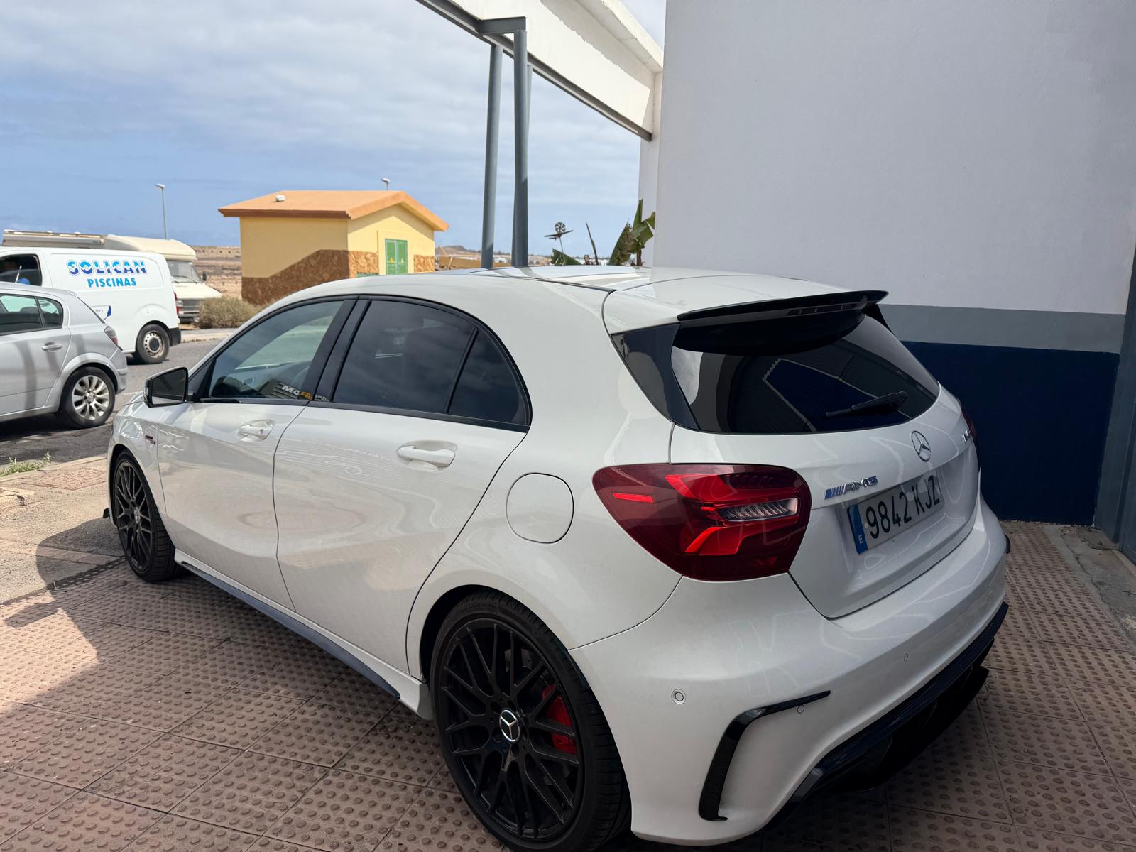Mercedes-Benz A45 AMG blanco estacionado, con llantas de aleación negras y detalles deportivos. Se observa un entorno urbano con otros vehículos y un edificio al fondo.