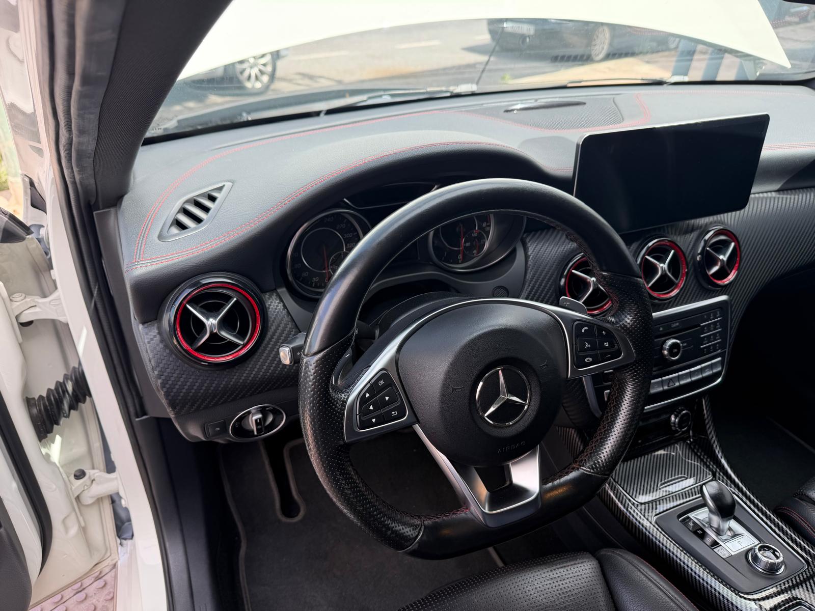 Interior de un automóvil Mercedes-Benz, mostrando el volante de cuero, pantalla central y detalles en fibra de carbono. Se aprecian los aireadores con borde rojo y el diseño moderno del tablero.