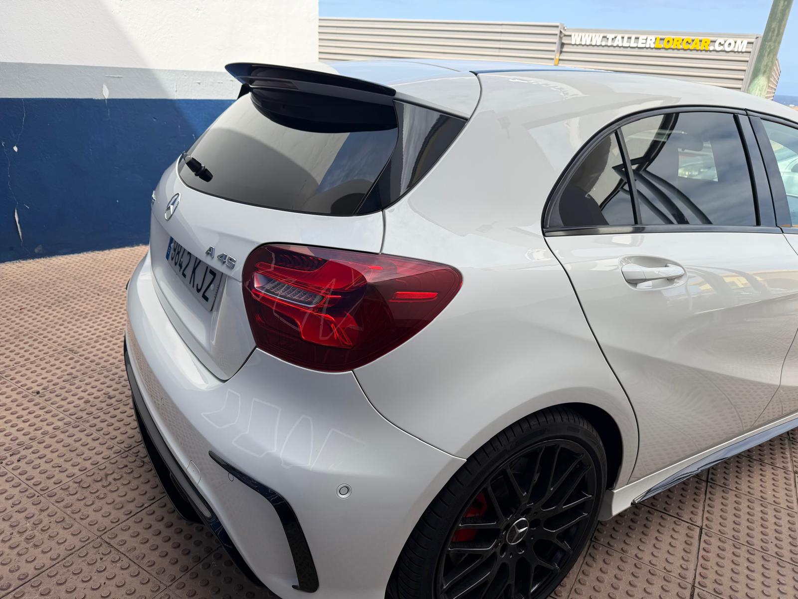 Mercedes-Benz A45 en color blanco, vista trasera con alerón y llantas negras de alto rendimiento, estacionado en un taller mecánico.