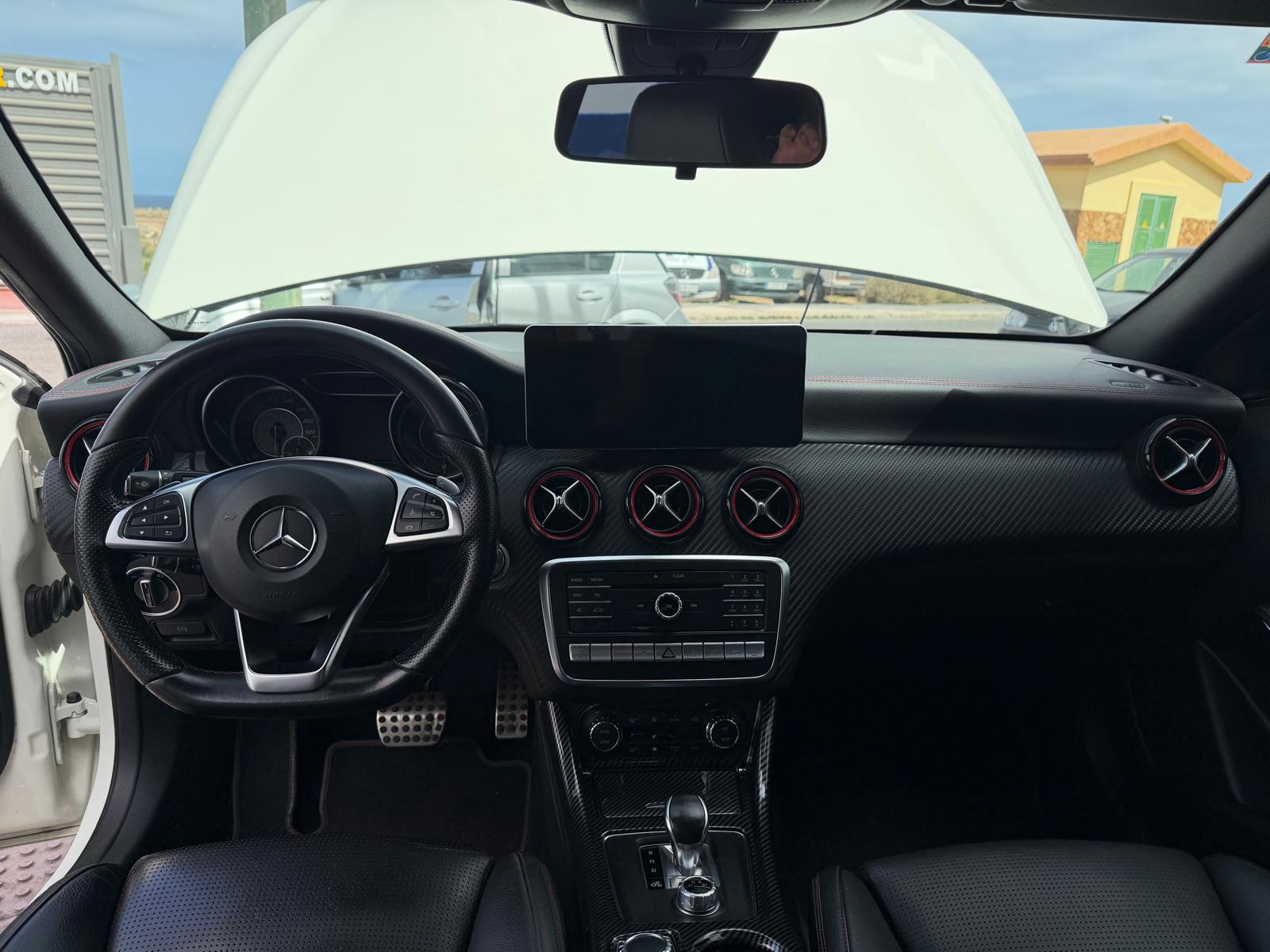 Interior de un automóvil Mercedes-Benz con volante deportivo, pantalla central y detalles en carbono. Se observa el capó levantado, lo que sugiere una revisión mecánica.