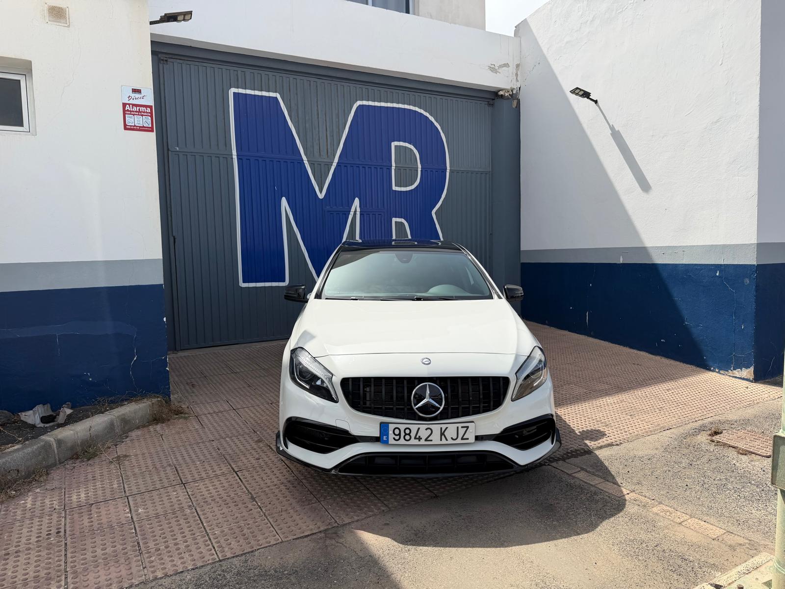 Mercedes-Benz blanco estacionado frente a un taller con la letra "MR" en la puerta. El vehículo tiene matrícula 9842 KJZ y se encuentra en un entorno urbano.